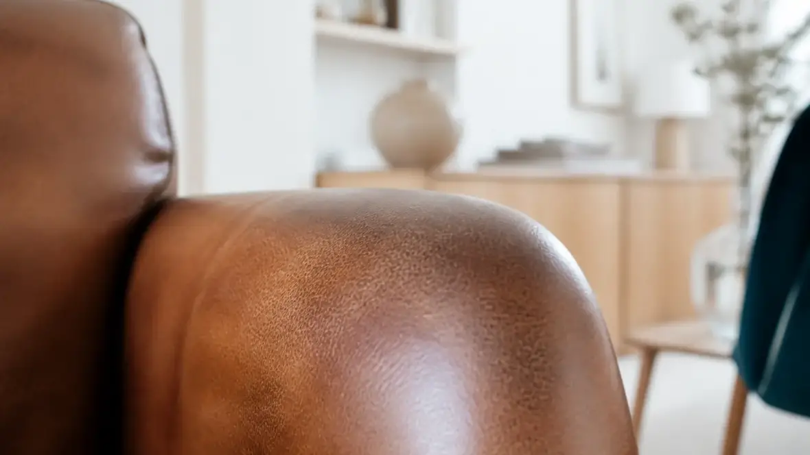 Gros plan sur un accoudoir de fauteuil en cuir brun, grain irrégulier et pores visibles, plus foncé là où la main se pose
