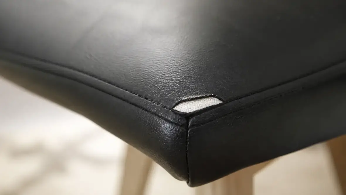 Coin d’une assise de chaise en simili cuir noir, couture ouverte laissant voir l’envers textile sous l’enduction