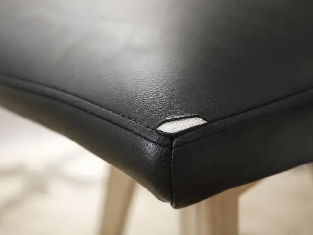 Coin d’une assise de chaise en simili cuir noir, couture ouverte laissant voir l’envers textile sous l’enduction